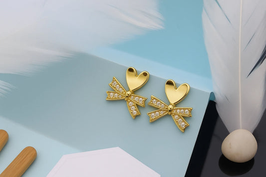 Gold bow-shaped earrings on a light blue surface with a white feather and wooden object.