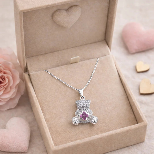 Silver teddy bear pendant necklace with purple gemstone in a beige jewelry box, surrounded by pink roses and heart-shaped objects.