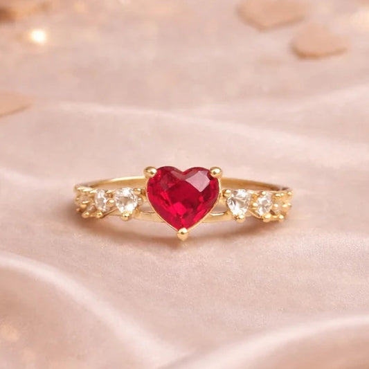 Gold ring with a red heart-shaped gemstone in a red velvet box on a soft fabric background.