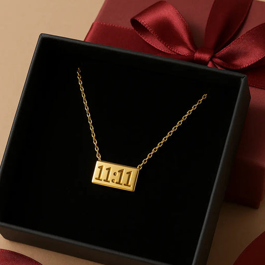Gold necklace with '11:11' pendant in a black box with a red ribbon on a beige background