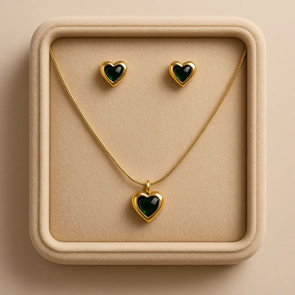 Gold heart-shaped necklace and earrings with green stones on a beige display box.