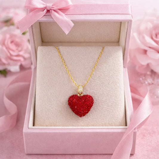 Red heart-shaped pendant necklace in a pink box with a bow, surrounded by pink flowers.