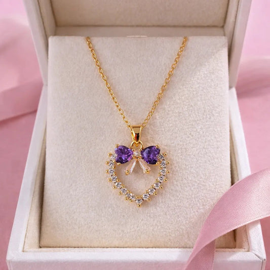 Gold necklace with a heart-shaped pendant featuring purple stones in a white box on a pink background