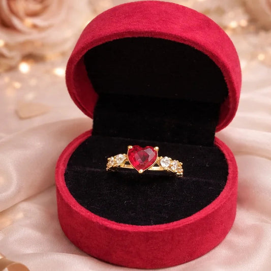 Gold ring with a red heart-shaped gemstone in a red velvet box on a soft fabric background.