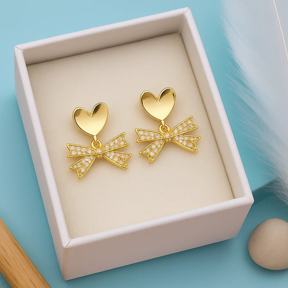 Gold heart-shaped earrings with bow details in a box on a light blue background
