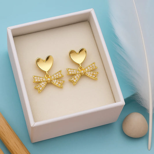 Gold heart-shaped earrings with bow details in a box on a light blue background