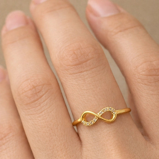 Gold infinity ring on a person's finger against a neutral background