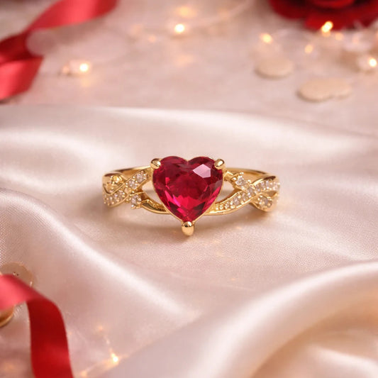 Gold ring with a red heart-shaped gemstone on a white fabric background with red ribbons and lights.