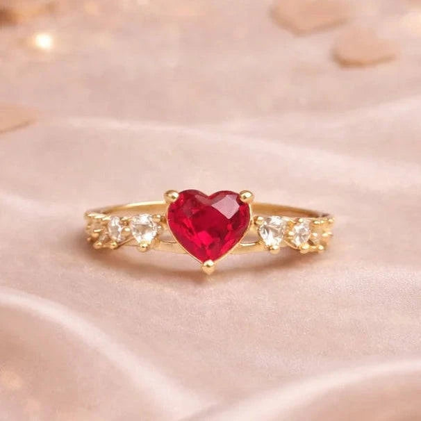 Gold ring with a red heart-shaped gemstone in a red velvet box on a soft fabric background.