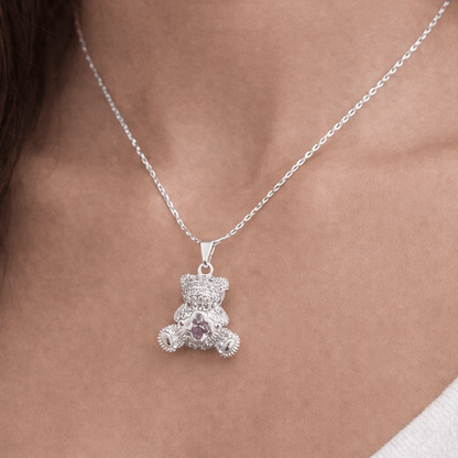 Silver teddy bear pendant necklace with a pink gemstone on a person's neck.