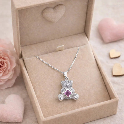 Silver teddy bear pendant necklace with purple gemstone in a beige jewelry box, surrounded by pink roses and heart-shaped objects.